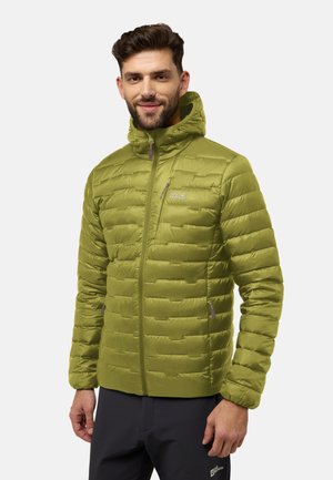 Mann lächelt, trägt eine grüne gesteppte Jacke mit Kapuze und schwarze Hose, steht vor einem einfarbigen hellen Hintergrund.