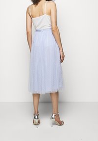 Kvinna klädd i en vit ärmlös topp, ljusblå genomskinlig midiklänning med prickar, och silverfärgade högklackade sandaler, stående mot en vit bakgrund.