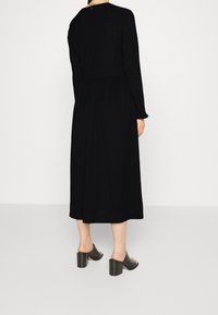 Mujer vestida con un largo vestido negro y sandalias de tacón negro tipo slip-on, de pie sobre un fondo blanco liso, mirando hacia atrás.