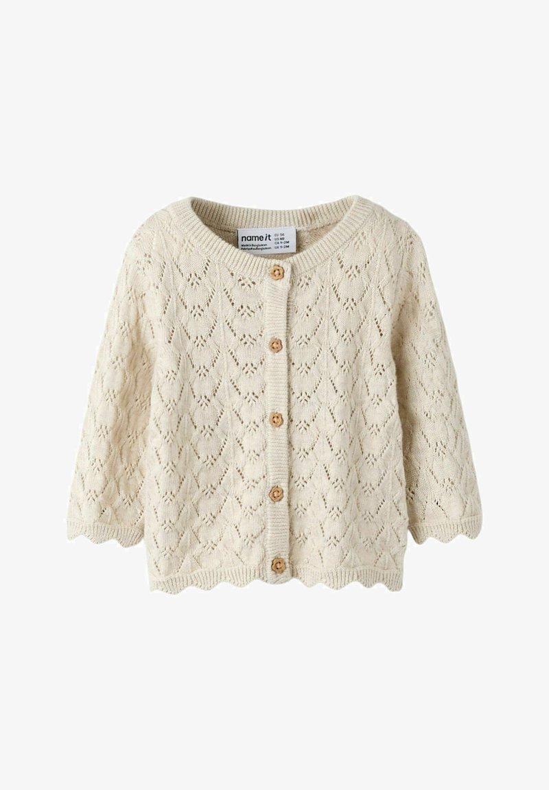 Cardigan bébé tricoté crème avec bords festonnés, boutons en bois, et motif losange en dentelle.