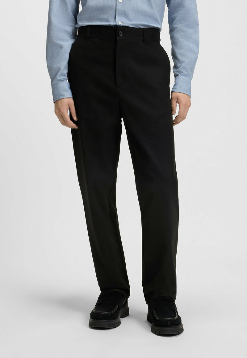 Pantaloni neri con una vestibilità dritta, dotati di chiusura a bottone, tasche laterali e una texture del tessuto liscia, abbinati a scarpe nere.