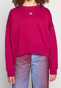 Fioletowa, przycięta bluza wykonana z miękkiego materiału, z długimi rękawami i białym logiem Adidas na piersi. Prosty, luźny krój.