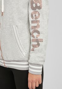 Manica di una felpa zip-up grigia chiara con testo metallico punteggiato, polsino a righe e mano rilassata accanto a pantaloni neri.