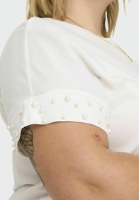 Hvid kortærmet top med perleaccent på manchetterne; glat tekstur, rund halsudskæring og synlig tatovering på armen.