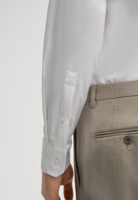 Camicia bianca con maniche lunghe e polsini con bottoni. Tessuto liscio, design aderente. Abbinata a pantaloni beige chiaro con bottone e tasca.