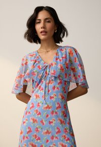 Florales Kleid aus hellblauem Stoff mit transparenten, mehrlagigen Volantärmeln und Bindeband am Ausschnitt. Verziert mit lebhaften rosa und orangenen Mustern.