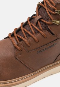 Bruine leren enkelboot met zwarte mesh accenten, ronde bruine veters, gouden oogjes en witte rubberen zool; bevat een logo aan de zijkant.
