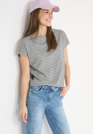 Femme souriante, portant un t-shirt rayé gris et blanc, un jean bleu clair et une casquette de baseball rose clair, debout contre un mur blanc.