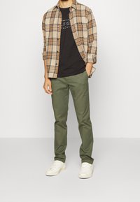 En rutiga skjorta i brunt och beige över en svart t-shirt, parat med olivgröna byxor och vita sneakers. Passformen är avslappnad.
