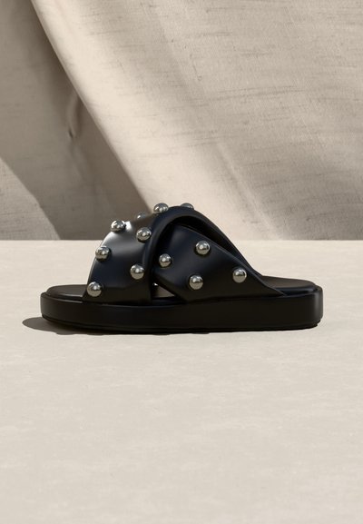 Sandale noire à glissière avec larges brides croisées décorées de clous métalliques ronds, présentée sur une surface et un fond beige clair.