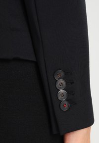 Mustan räätälöidyn pukujaketin hiha, jossa on sileä pinta, neljä tummaa nappia punaisella yksityiskohdalla ja muotoiltu istuvuus.