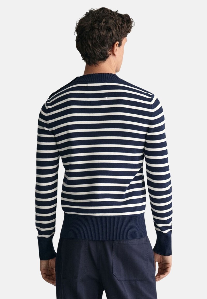 Hombre de espaldas vistiendo un suéter de rayas horizontales azul marino y blanco y pantalones azul marino oscuro sobre fondo blanco.