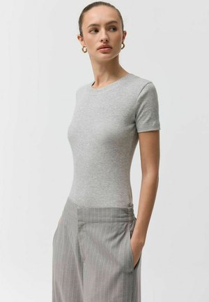 Kobieta ubrana w dopasowany szary krótki top z krótkim rękawem i wysokowistowe szare spodnie w prążki, ręce w kieszeniach, spoglądająca na bok.