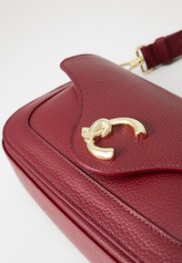 Burgundy textured leather shoulder bag with curved flap and gold-tone metal clasp, featuring an adjustable matching strap.