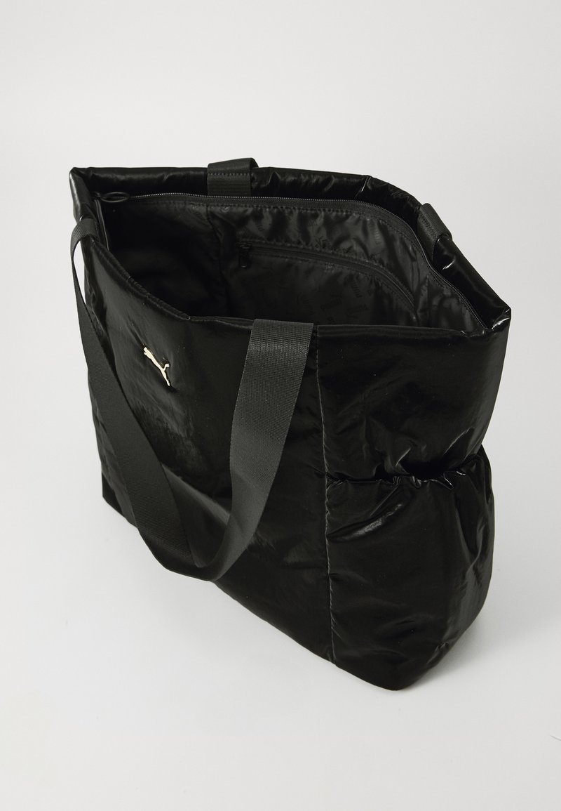 Bolso tote acolchado negro con acabado brillante, asas dobles en la parte superior y bolsillo interior. Presenta un prominente logo dorado en un lado.