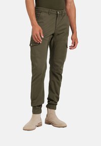 Pantaloni cargo verde oliva con polsini elasticizzati, multiple tasche, tessuto testurizzato e silhouette aderente, abbinati a stivaletti beige alla caviglia.