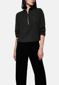 Zwart topje met ritssluiting, hoge kraag, raglanmouwen en een losse pasvorm. Gecombineerd met zwarte wijde broek met strepen, met een zachte textuur.