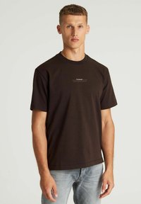 Bruine katoenen t-shirt met korte mouwen, ronde hals en een klein wit logo op de borst. Gecombineerd met lichtblauwe denim jeans.