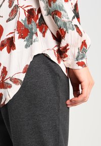 Floral blouse with red and green patterns on a cream background, paired with dark gray tapered pants; features a buttoned cuff and textured fabric.