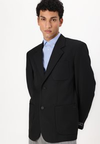 Μαύρο σακάκι κομμένο με δύο μπροστινές τσέπες, με μανσέτες και τρία κουμπιά. Φορεμένο πάνω από ένα ανοιχτό μπλε πουκάμισο με γιακά. Μοναδική υφή υφάσματος.