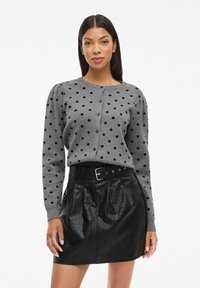 Grå stickad cardigan med svarta prickar, rund nacke, knappstängning och puffärmar, i kombination med en glänsande svart texturerad kjol.