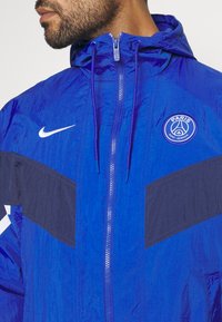 Man bär en blå Paris Saint-Germain Nike-jacka med dragkedja fram och luva, visar bröstet och nedre delen av ansiktet.