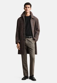 Hnedý károvaný plášť s veľkým golierom, čierny top a béžové nohavice. Má tmavé gombíky a textúrovaný povrch látky. Čierne topánky dopĺňajú outfit.