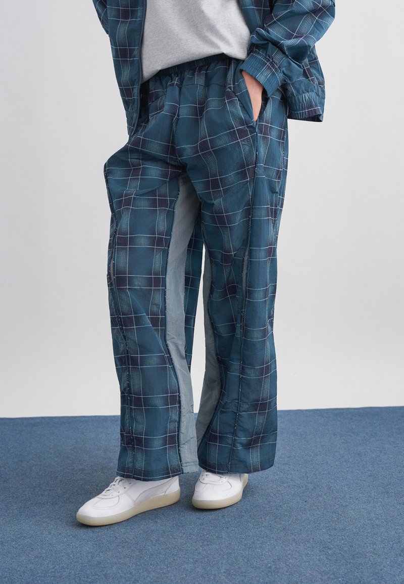 Άτομο με μπλε καρό φαρδύ παντελόνι και ταιριαστό σακάκι, φορώντας λευκά αθλητικά παπούτσια, στέκεται σε μπλε μοκέτα.