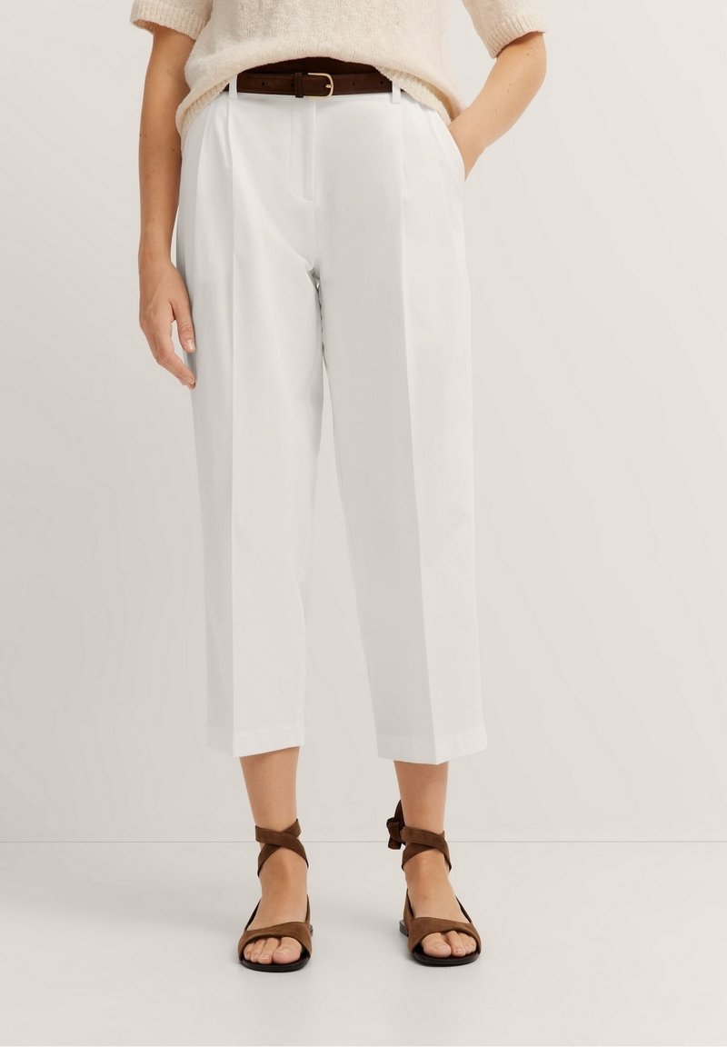 Femme portant un pantalon court blanc avec pli devant, ceinture marron, pull beige à manches courtes et sandales plates marron à lanières autour du pied.