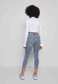 Top corto de manga larga blanco con puños con volantes, combinado con jeans ajustados de color azul claro que tienen dos bolsillos traseros y un parche de cuero marrón en la cinturilla.