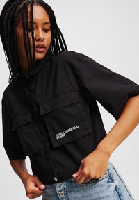 Čierna skrátená bunda s krátkymi rukávmi, dvoma prednými vreckami a bielym logom "KARL LAGERFELD JEANS". Vyrobená z bavlnenej tkaniny s hladkou textúrou.