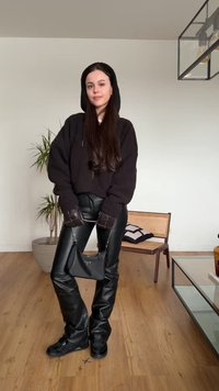 Joven mujer con sudadera con capucha negra, pantalones de cuero, guantes y zapatillas deportivas, sostiene un bolso negro y está de pie sobre un suelo de madera en una habitación moderna.