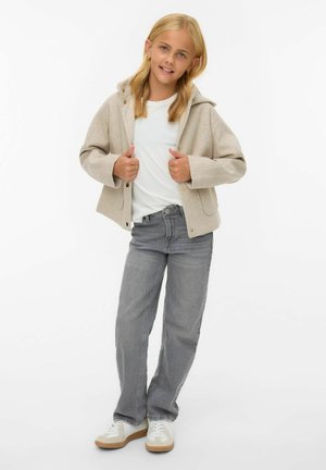 Veste à capuche beige en tissu doux, t-shirt blanc en dessous, jeans gris et baskets blanches avec des semelles beiges. Fond uni.