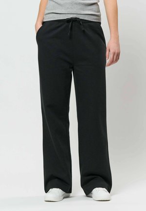 Personne portant un pantalon de survêtement noir ample avec cordon à la taille, chemise grise rentrée, mains dans les poches et baskets blanches sur fond uni.