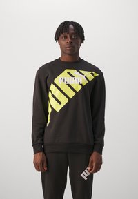 Felpa nera con un grande logo geometrico giallo e testo bianco. Polsini e orlo a costine con pantaloni abbinati visibili nell'immagine.