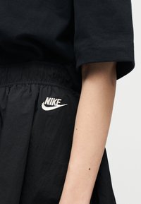 Κοντινό πλάνο ενός ατόμου που φοράει μαύρο σορτς Nike με ελαστική μέση και λευκό λογότυπο Nike, μαζί με μέρος του μανικιού μαύρου πουκαμίσου και του αντιβραχίου.