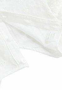 Blouse en coton blanc pour bébé avec broderies florales, présentant un col rabattu et des fermetures à boutons. Étiquette visible avec "NEXT baby."