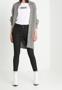 Grå stickad cardigan, vit logot-tröja, svarta rutiga slim-fit jeans och blanka vita ankelboots. Fokusera på material- och texturdetaljer.