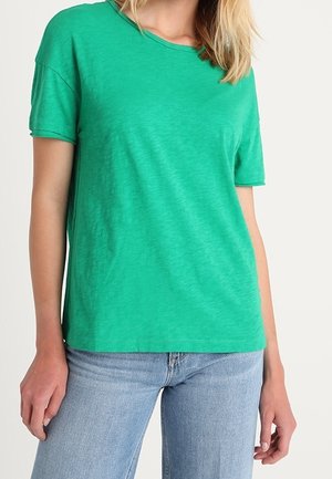 Kobieta w prostym zielonym t-shircie z krótkim rękawem, ubrana w jasnoniebieskie dżinsy, stojąca na białym tle.