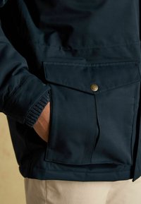 Tmavomodrá bunda s gombíkovou náprsní kapsou a rebrovanými manžetami. Materiál vyzerá hladký s jemným leskom. Výstrih je klasického štýlu.