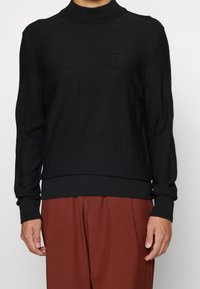 Pull en maille noir avec un col roulé côtelé et des poignets. Caractérisé par un logo ton sur ton sur la poitrine. Associe à un pantalon marron à coupe ample.
