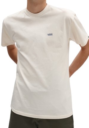 Man draagt een effen off-white Vans T-shirt met een klein marineblauw logo op de borst en donkere broek, handen achter de rug.