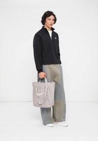 Γκρίζα τσάντα ώμου Nike με λογότυπο, από νάιλον υλικό, διπλές λαβές. Το μοντέλο φοράει ένα μαύρο πουλόβερ με φερμουάρ στον λαιμό και γκρι κλασικά παντελόνια.