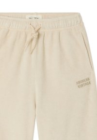 Pantalon de survêtement en éponge beige avec taille élastique, cordon de serrage, poches latérales, et « American Vintage » brodé sur la cuisse droite.