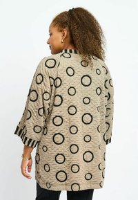 Texturierte beiger Jacke mit schwarzen Kreis-Mustern, Dreiviertelärmeln und gestreiften Akzenten an den Bündchen. Knopfdetail an der Seite.