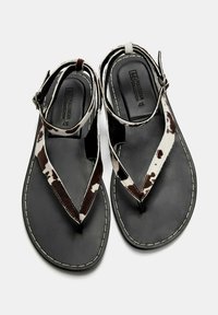 Sandali in pelle di mucca marroni e bianchi con suola nera, dotati di un design a T e cinturino alla caviglia regolabile. Dettagli di cucitura a contrasto.