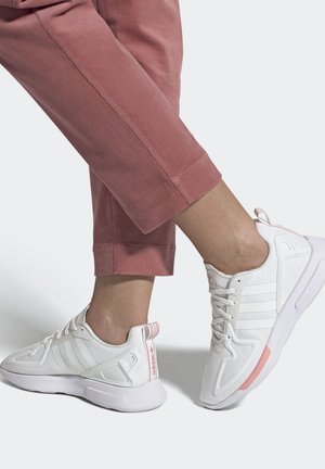 Stopy w białych butach Adidas z jasnoróżowymi akcentami, w zestawieniu z krótkimi różowymi spodniami na jednolitym tle.