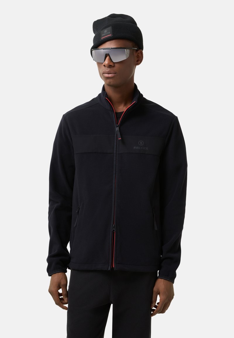 Black fleece jacket with zip front, red accents, and two side pockets. Paired with a black beanie and mirrored sunglasses.