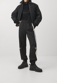 Giacca bomber nera con texture trapuntata e zip frontale, abbinata a pantaloni jogger neri con accenti grigi e strisce laterali, indossati con sneakers nere.