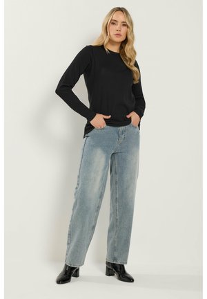 Ung kvinde med langt blondt hår iført en sort langærmet top, lyseblå jeans med vide ben og sorte støvler med hæl, stående med hænderne i lommerne.